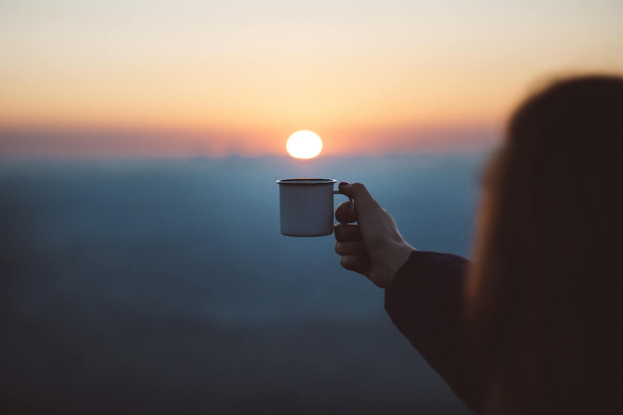 A woman holding a cup of coffee against a stunning sunrise backdrop, creating a serene and cozy atmosphere.